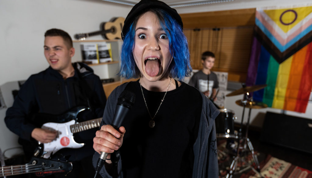Jente med blått hår holder mikrofon og gjeiper til kamera, med gitarist og trommis i bakgrunnen.